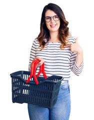 Beautiful young brunette woman holding supermarket shopping basket pointing finger to one self smiling happy and proud
