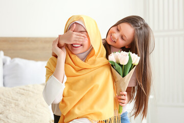 Little girl with tulips closing her Muslim mother's eyes in bedroom