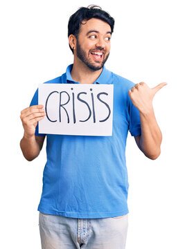 Young hispanic man holding crisis message paper pointing thumb up to the side smiling happy with open mouth