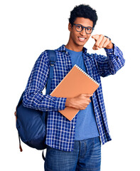 Young african american man wearing student backpack holding book pointing to you and the camera...