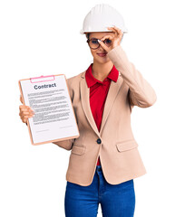 Young beautiful woman wearing architect hardhat holding clipboard with contract document smiling happy doing ok sign with hand on eye looking through fingers