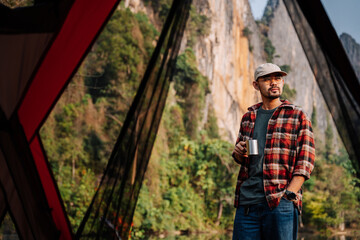 Fototapeta na wymiar Asian man standing in front of a tent holding a hot drink mug. coffee and camping.