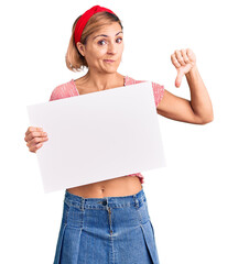 Young blonde woman holding blank empty banner with angry face, negative sign showing dislike with thumbs down, rejection concept