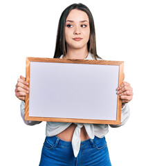Young hispanic woman holding empty white chalkboard smiling looking to the side and staring away thinking.