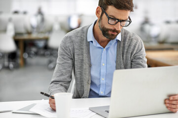 Hes hard at work on his laptop. A young businessman going over paperowrk while working on his...