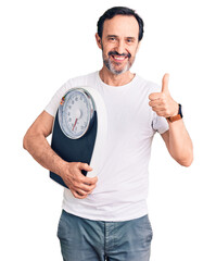 Middle age handsome man holding weighing machine smiling happy and positive, thumb up doing excellent and approval sign