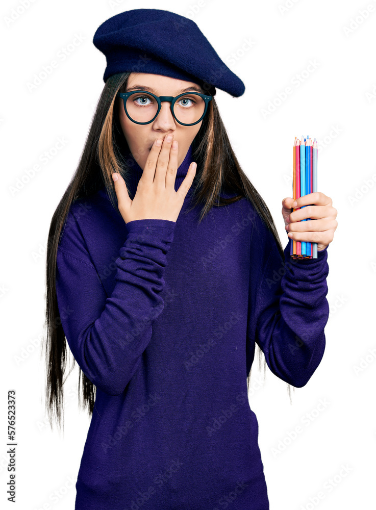 Wall mural Young brunette girl with long hair holding colored pencils covering mouth with hand, shocked and afraid for mistake. surprised expression