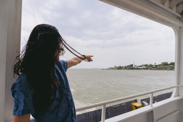 Young Latin man with long hair pointing to land from the deck of a ferry while the wind ruffles his...