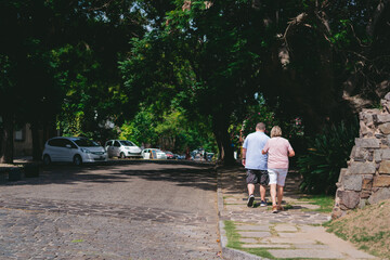 Elderly couple walking along the sidewalk of a cobblestone street with a tunnel of trees. Copy space.
