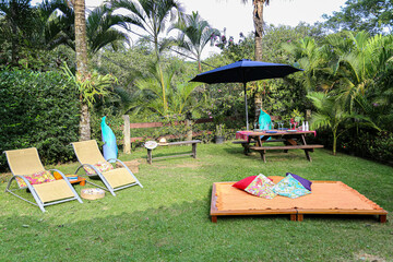 belo espaço ao ar livre em estilo zen para relaxamento, meditação e lazer com espreguiçadeiras, almofadas, mesa de piquenique de madeira e umbrelone em meio à vegetação tropical.