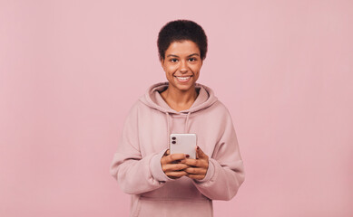 Smiling attractive black girl holding smartphone in her hands and looking at camera. Portrait of cheerful young African woman wearing casual hoodie and standing over pink backdrop 