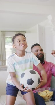 Vertical video of african american father and son watching match and supporting, in slow motion
