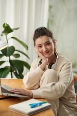 Portrait of smiling college student sitting at desk with opened laptop and looking at camera