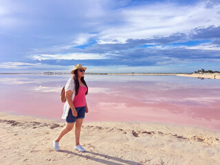 Latin adult woman with shorts, pink shirt, hat and sunglasses walks on the sand next to the pink colored lagoon with a high concentration of salt, Las Coloradas in Yucatan Mexico