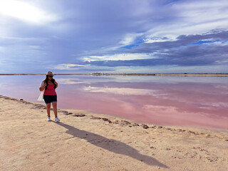 Latin adult woman with shorts, pink shirt, hat and sunglasses walks on the sand next to the pink colored lagoon with a high concentration of salt, Las Coloradas in Yucatan Mexico