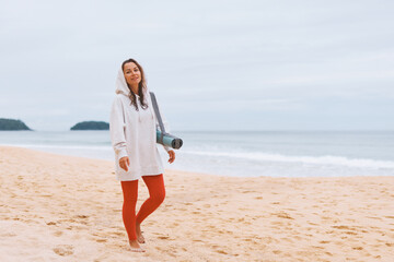 Young woman white hoodie, walking with yoga mat, strolling along seashore in early hours of morning