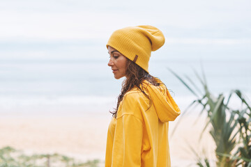 Young woman in yellow hoodie, yellow hat standing on seashore in a relaxed state. Tropical view - 576553103