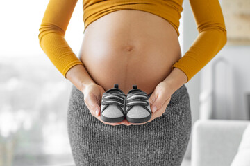 Pregnant woman expecting baby boy holding dark blue shoes in gender reveal during pregnancy. Expectant mother showing baby bump belly. Shopping baby clothing and gender reveal concept - Powered by Adobe