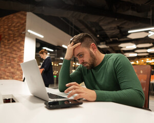 Bearded man holding his head while working on a laptop in an open space office. Red-haired caucasian woman uses smartphone. 