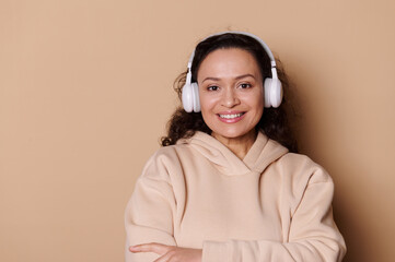 Close-up portrait of middle-aged beautiful multi-ethnic woman, with wireless headphones, smiling a toothy smile looking at camera, isolated beige background. Pretty female listens podcast, sound track