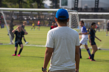 Father standing and watching his daughter playing football in a school tournament on a clear sky...