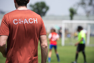 Fototapeta premium Male youth football coach standing and watching his team playing in a school tournament on a clear sky and sunny day. Sport, active lifestyle, happy activity and coaching concept. 