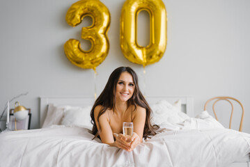 Obraz na płótnie Canvas Happy young woman meets her thirtieth birthday in bed, holding a glass of champagne in her hands and smiling
