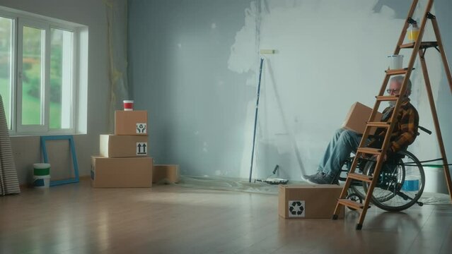 An elderly disabled man moves in a wheelchair with a cardboard box. The pensioner puts the box looks around and plans repairs. Room with window, stepladder, wallpaper rolls, roller and paint.