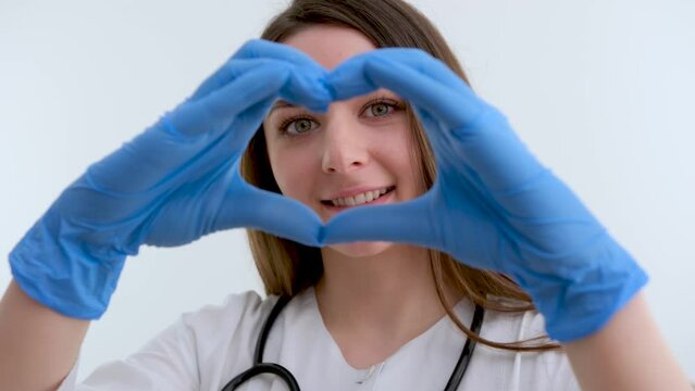Doctor hands in gloves in the heart shape against the background of his body face and medical gown. closeup. Female hand in blue medical gloves show heart sign. High quality photo