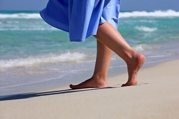 Barefoot woman in blue dress walking by the sand on sea waves background. Female legs on a coast, beach vacation - obrazy, fototapety, plakaty