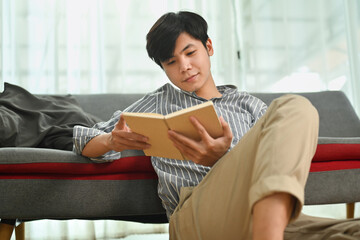 Young asian man sitting on floor and reading book, spending leisure weekend tine at home. Leisure and people concept