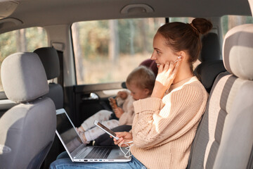 Profile portrait of woman wearing beige jumper sitting with her baby daughter in safety chair on backseat of car, holding mobile phone in hands, listening music, having video call.