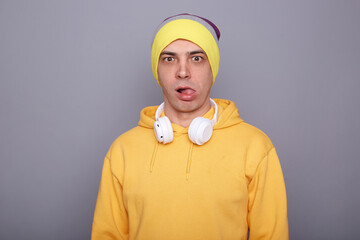 Portrait of childish palyful man wearing yellow hoodie, beanie hat and headphones standing isolated over gray background, looking at camera with surprised expression, showing tongue out.