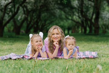 Fototapeta premium Mom and her daughters are lying on the blanket in a summer park. The family has fun outdoors