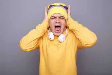 Portrait of shocked annoyed man wearing yellow hoodie, beanie hat and headphones, covering ears...