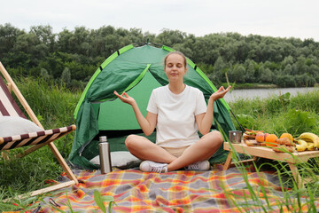Naklejka na ściany i meble Portrait of peaceful relaxed woman with brown hair wearing white t shirt, practicing yoga in open air while having camping, sitting near the tent by the water on ground.