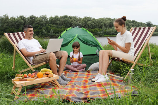 Portrait Of Busy Family, Mother, Father And Their Daughter Spending Time Near The River, Sitting On Deck-chairs Near The Tent, Husband Doing His Online Work, Wife Using Smart Phone, Kid Eating Fruit.