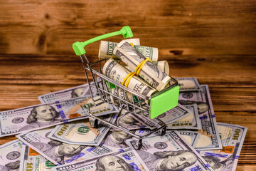 Small shopping cart with rolled up one hundred dollar banknotes on a wooden background