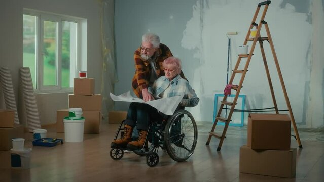An elderly man and woman in a wheelchair look through a sheet with an apartment project and communicate. An elderly couple is planning repairs in the house.
