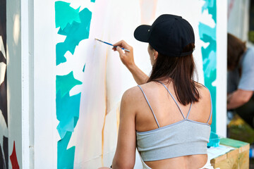 Girl artist draws with paint brush surreal woman portrait on white canvas at outdoor art painting festival, rear view, paintings art picture process. Woman paints atmospheric surreal picture