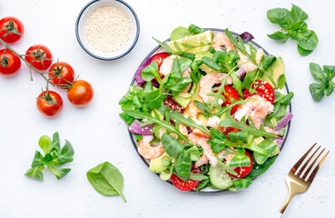 Delicious shrimp salad with avocado, cherry tomatoes, arugula, lamb lettuce, cucumber, red onion and sesame seeds on white table background. Top view