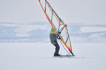 Fototapeta na wymiar A middle-aged man, a snowsurfer, rides a sailboard on a snow-covered frozen lake. Snowsurfing on a cloudy winter day.