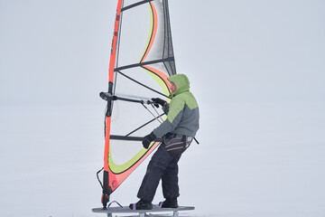 A middle-aged man, a snowsurfer, rides a sailboard on a snow-covered frozen lake. Snowsurfing on a cloudy winter day.