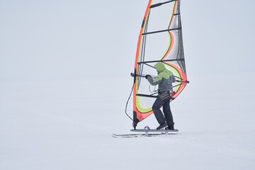 A middle-aged man, a snowsurfer, rides a sailboard on a snow-covered frozen lake. Snowsurfing on a cloudy winter day.