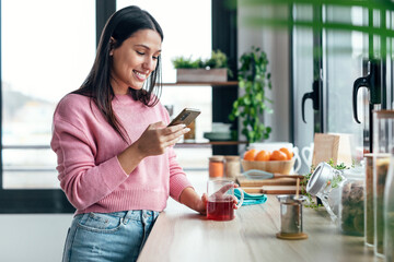 Beautiful woman using her mobile phone while drinking a cup of tea in the kitchen at home.