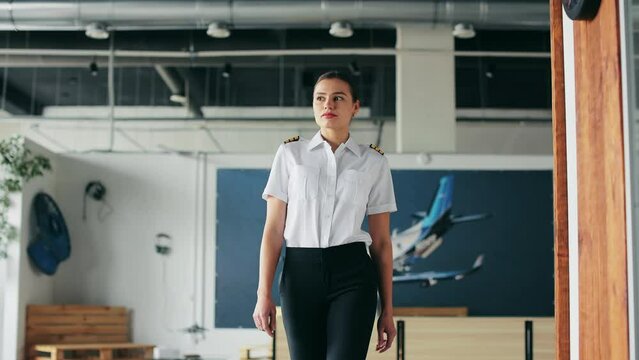Front view of serious female instructor, pilot teaching aviation. Beautiful, young woman wearing uniform, walking in classroom, looking aside. Concept of airplane and aviation.