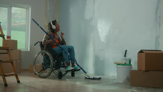 An African American woman in white headphones on a wheelchair paints a wall with white paint using a long roller. A young black woman with a disability makes repairs in room, enjoys music and sings.
