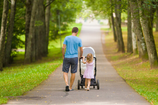 Young adult father and little daughter pushing white baby stroller and walking at town park in summer day. Spending time together and breathing fresh air. Enjoying stroll. Two child dad. Back view.