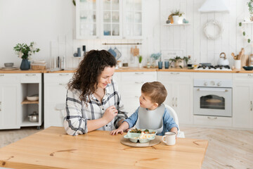 Young beautiful mother feeds her adorable toddler son with healthy vegetables and a chicken cutlet in a bright cozy scandinavian style kitchen. Child's lunch. Mock up, copy space
