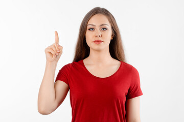 Happy lovely girl points finger up, shows advertisement on blank white space. Young brunette female stands over white background